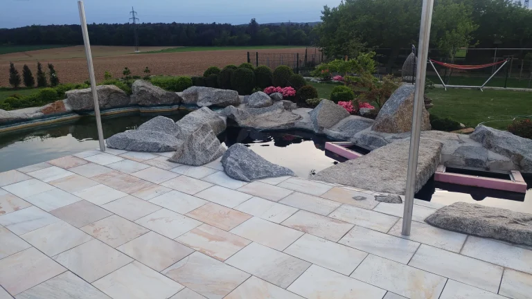Künstlicher Gartenteich, eingefasst mit Naturstein-Findlingen und umgeben von Naturstein-Bodenplatten.